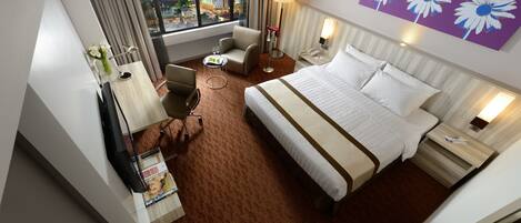 A hotel room with a large bed, a desk with a lamp, a chair, and a view of a cityscape through a large window.