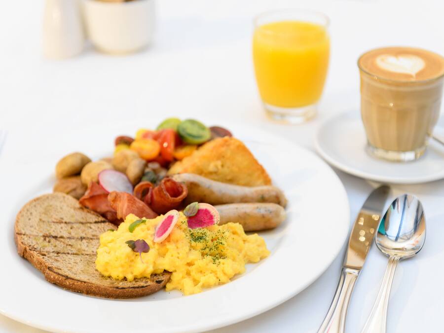 A breakfast plate with scrambled eggs, bacon, sausages, and toast, accompanied by a glass of orange juice and a cup of coffee.