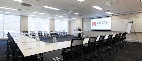 A conference room with a long table, black chairs, and a projector screen displaying 'MGM Macquarie University'.