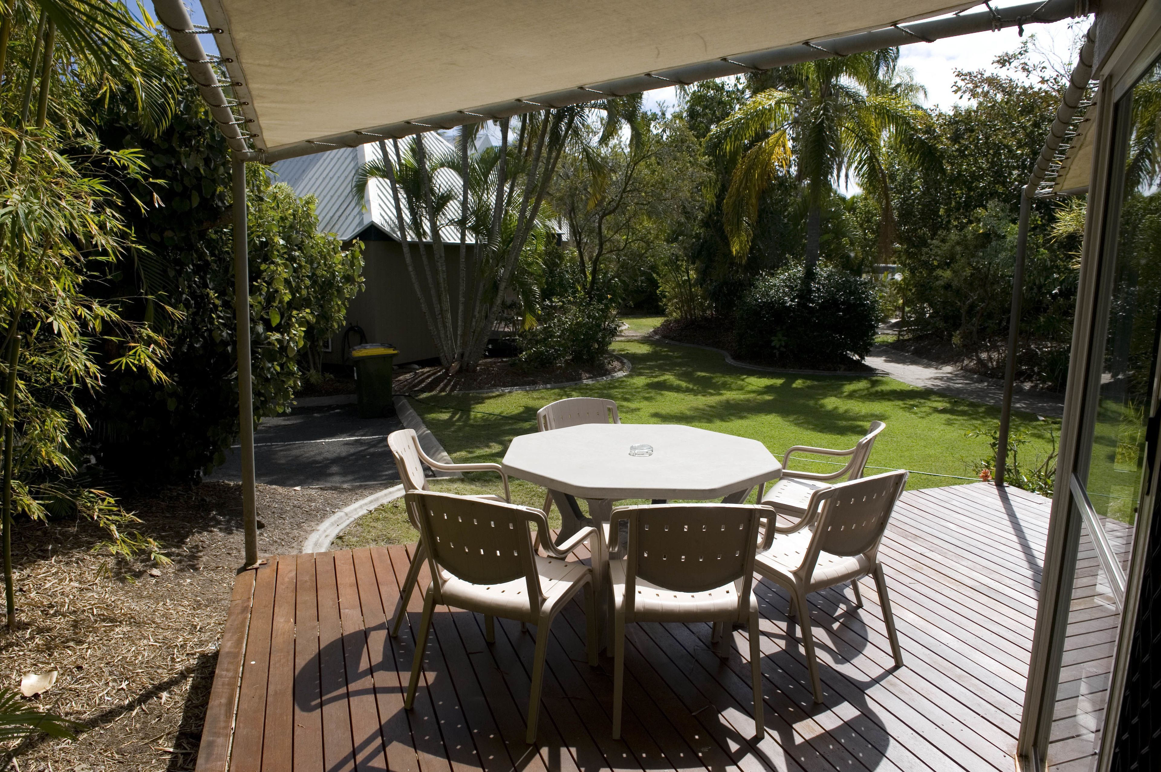 Un patio exterior con mesa y sillas, rodeado de un jardín con diversas plantas y árboles.