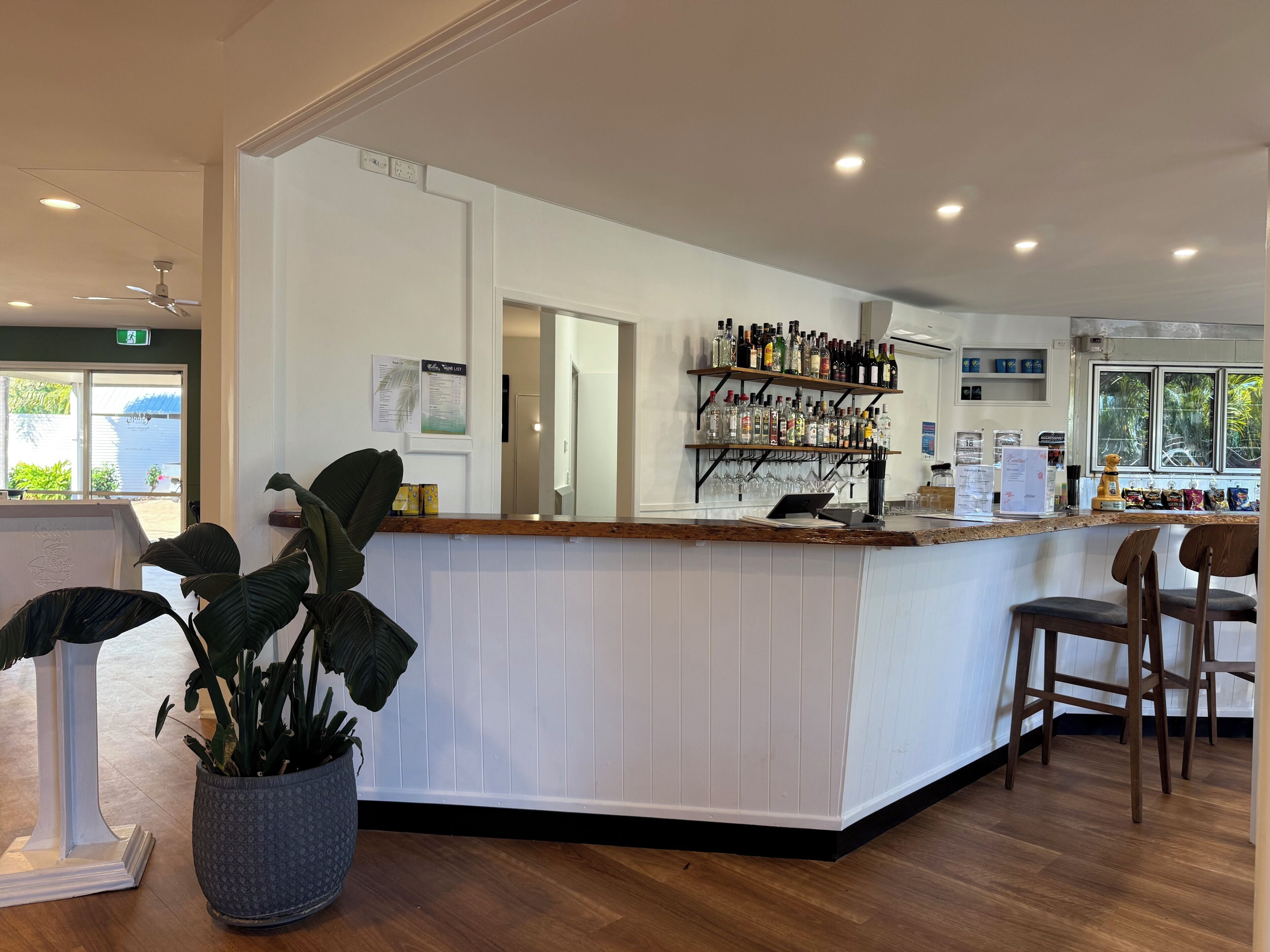 A bar area with a wooden counter, shelves with bottles, a laptop, and a potted plant.