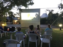Proyección de cine al aire libre con una pantalla grande, sillas de plástico blanco y altavoces sobre una mesa.