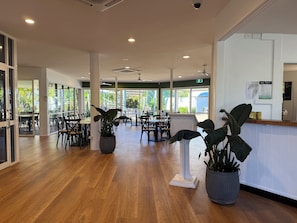 A spacious indoor area with wooden flooring, multiple tables and chairs, potted plants, and large windows allowing natural light. - Kellys Beach Resort (Bargara)