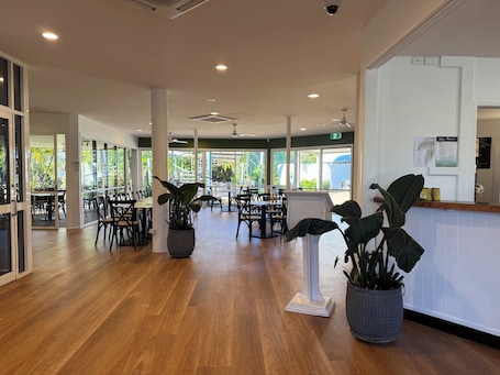 Um espaço amplo e interno com piso de madeira, várias mesas e cadeiras, plantas em vasos e janelas grandes que permitem a entrada de luz natural.. Kellys Beach Resort