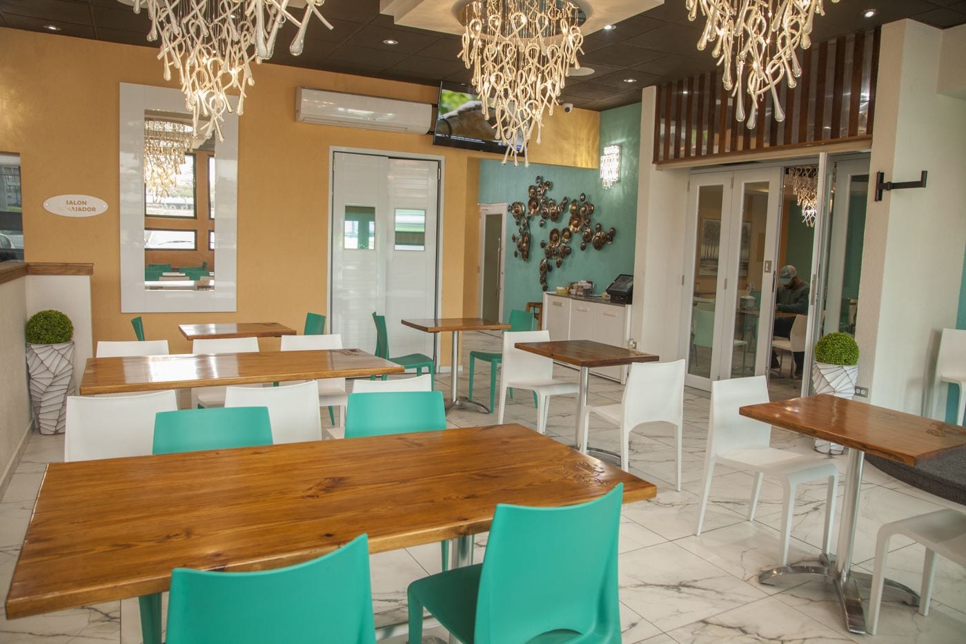 Un café moderne avec des tables en bois et des chaises de couleur turquoise, un téléviseur fixé au mur et des œuvres d’art décoratives.