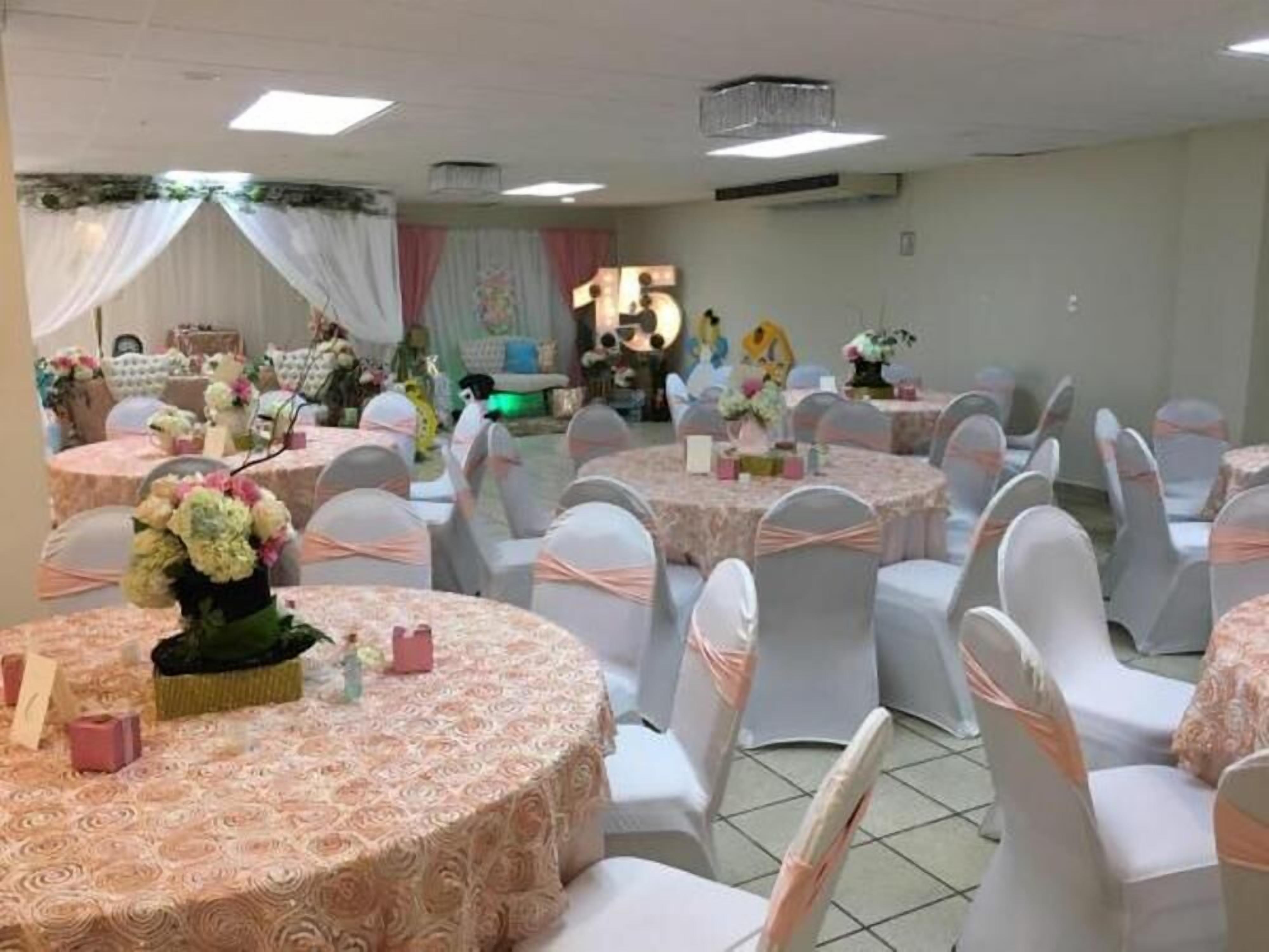 A banquet hall with round tables covered in floral tablecloths, white chairs with pink sashes, and a stage area with a backdrop.