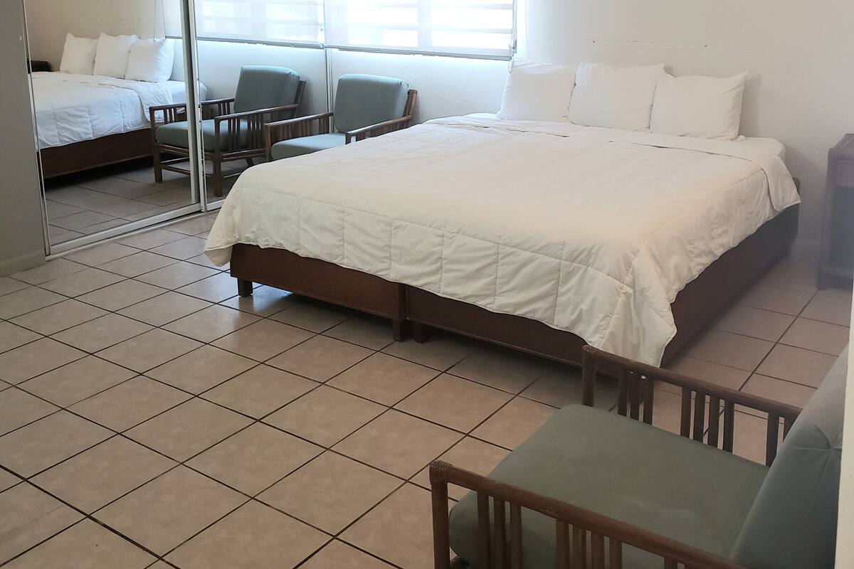 Une chambre d’hôtel avec un grand lit, un bureau, deux chaises et une porte-fenêtre coulissante donnant sur une autre pièce.