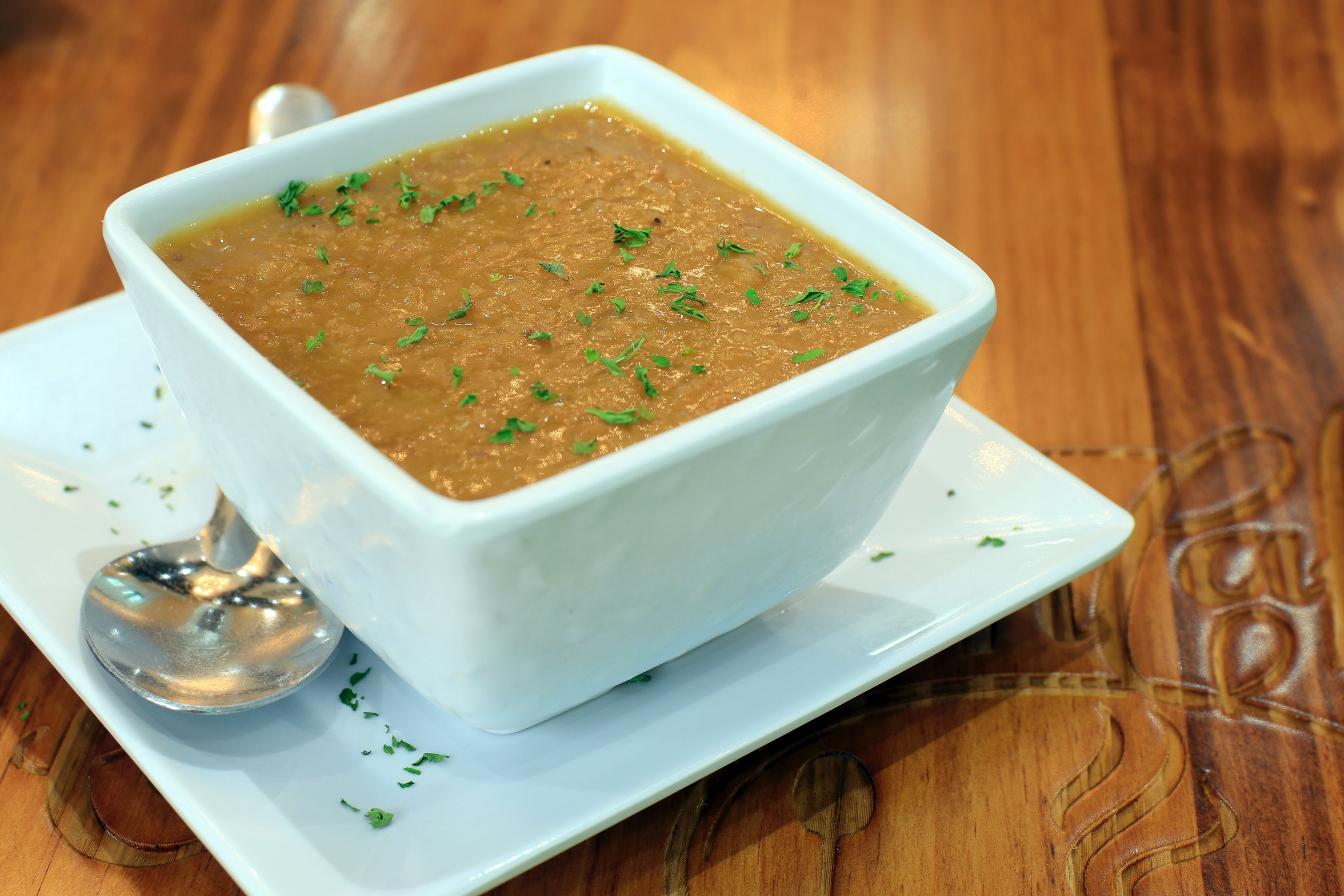 Un tazón blanco de sopa con una cuchara sobre un plato blanco, sobre una mesa de madera.