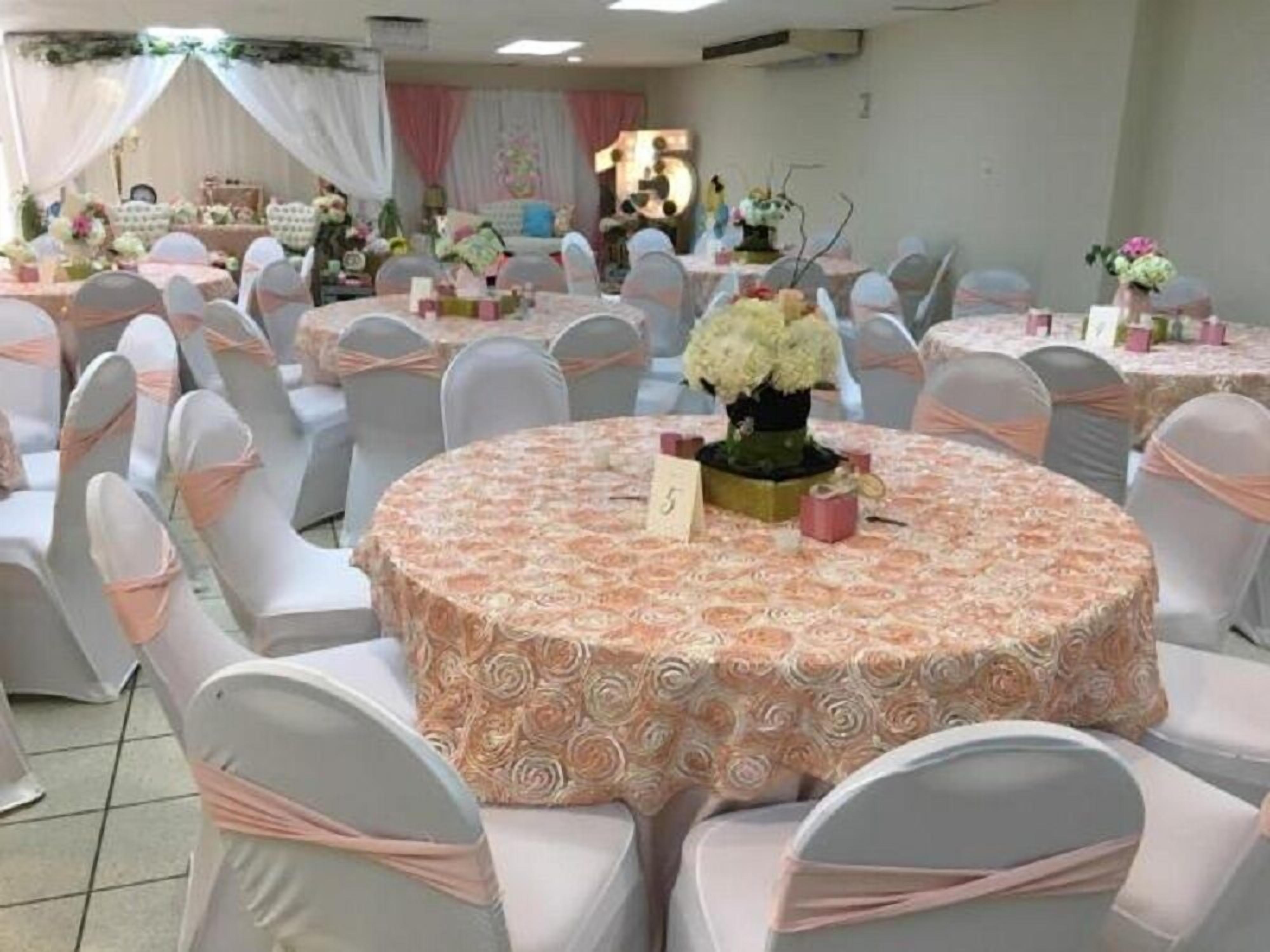 A banquet hall with round tables covered in floral tablecloths, chairs with white and pink sashes, and a stage area in the background.