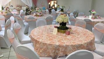 A banquet hall with round tables covered in floral tablecloths, chairs with white and pink sashes, and a stage area in the background.