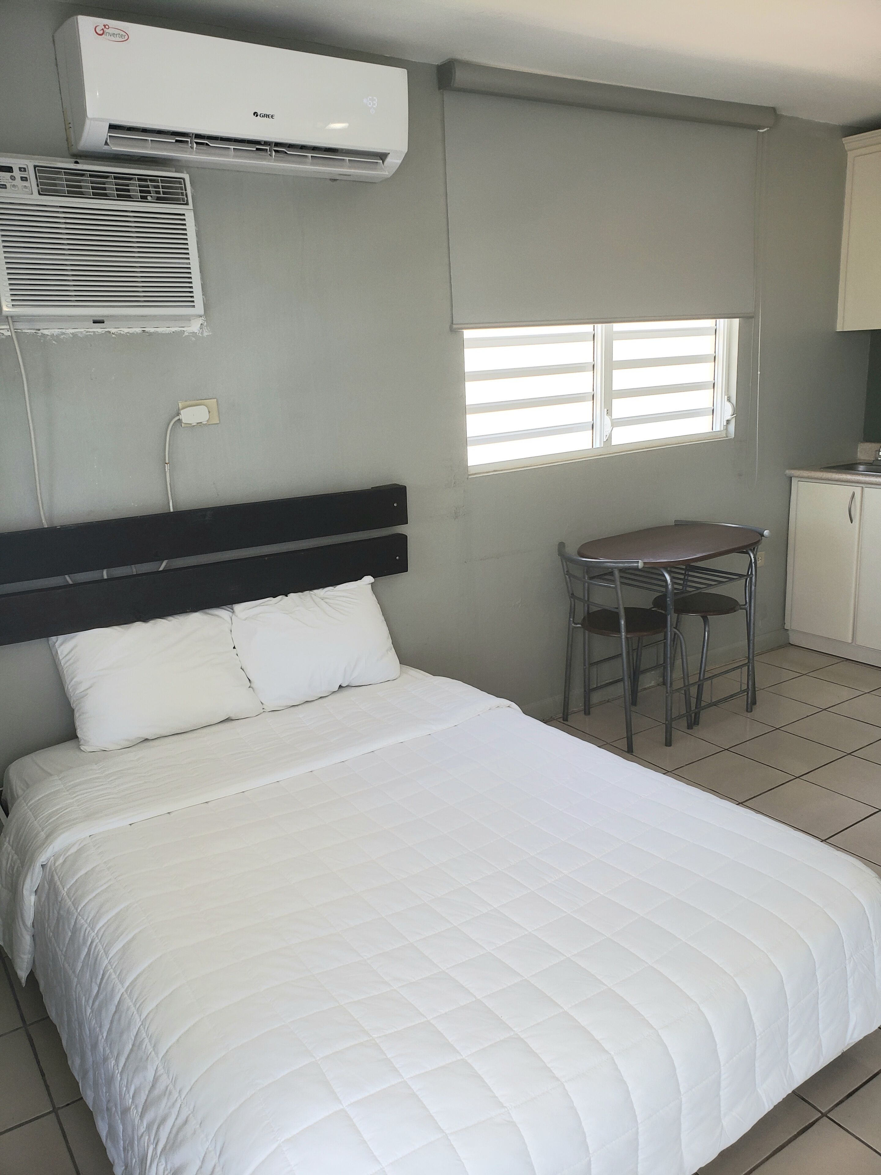 A hotel room with a bed, a small table with chairs, an air conditioning unit, and a window with blinds.