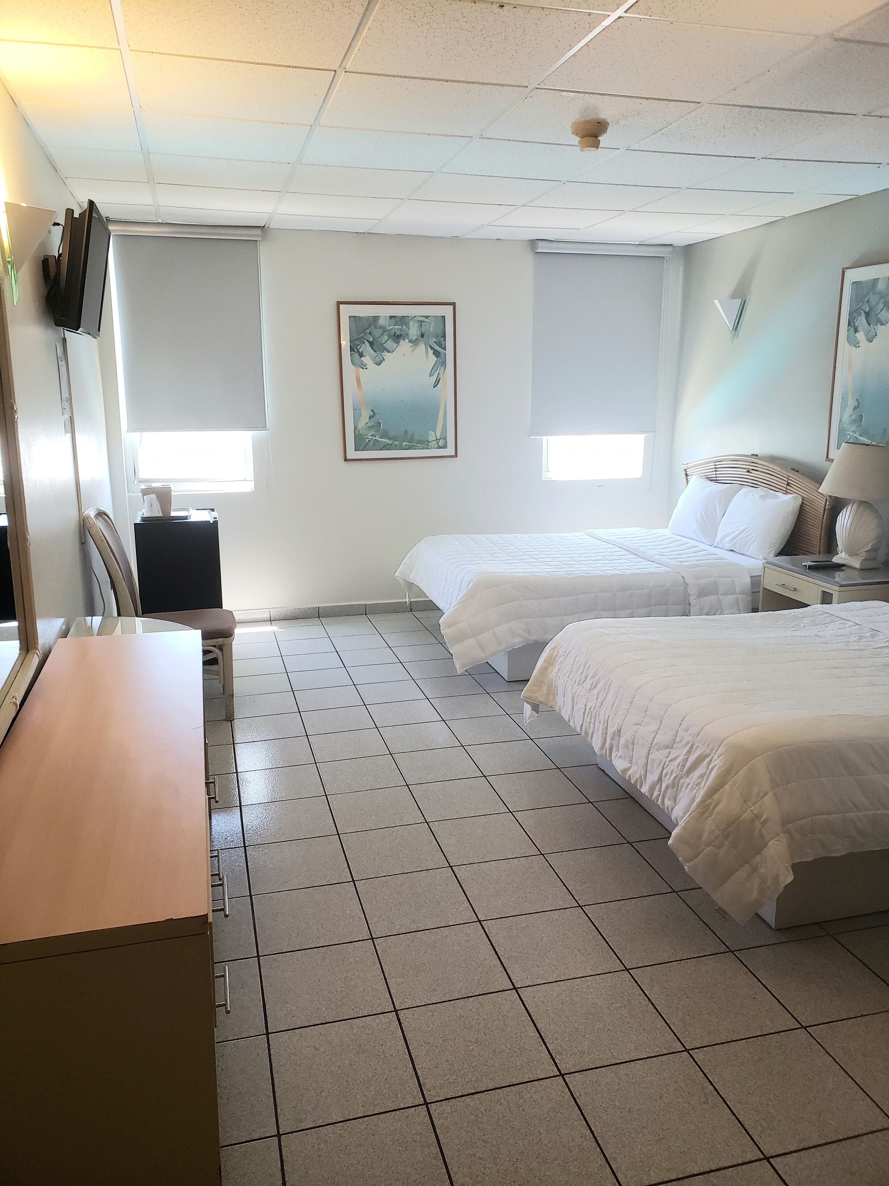 A hotel room with two beds, a desk, and a chair. There are two windows with blinds, a ceiling fan, and a painting on the wall.