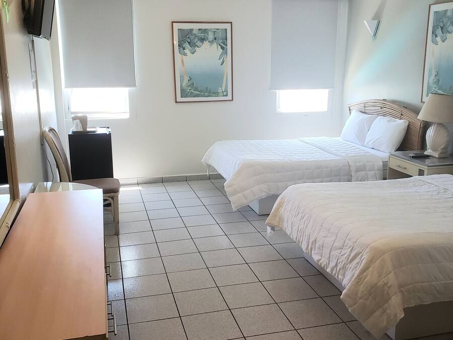 A hotel room with two beds, a desk, and a chair. There are two windows with blinds, a ceiling fan, and a painting on the wall.