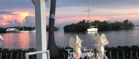 Mesa para dos con velas y un frutero, con vista a un cuerpo de agua con barcos y una puesta de sol.