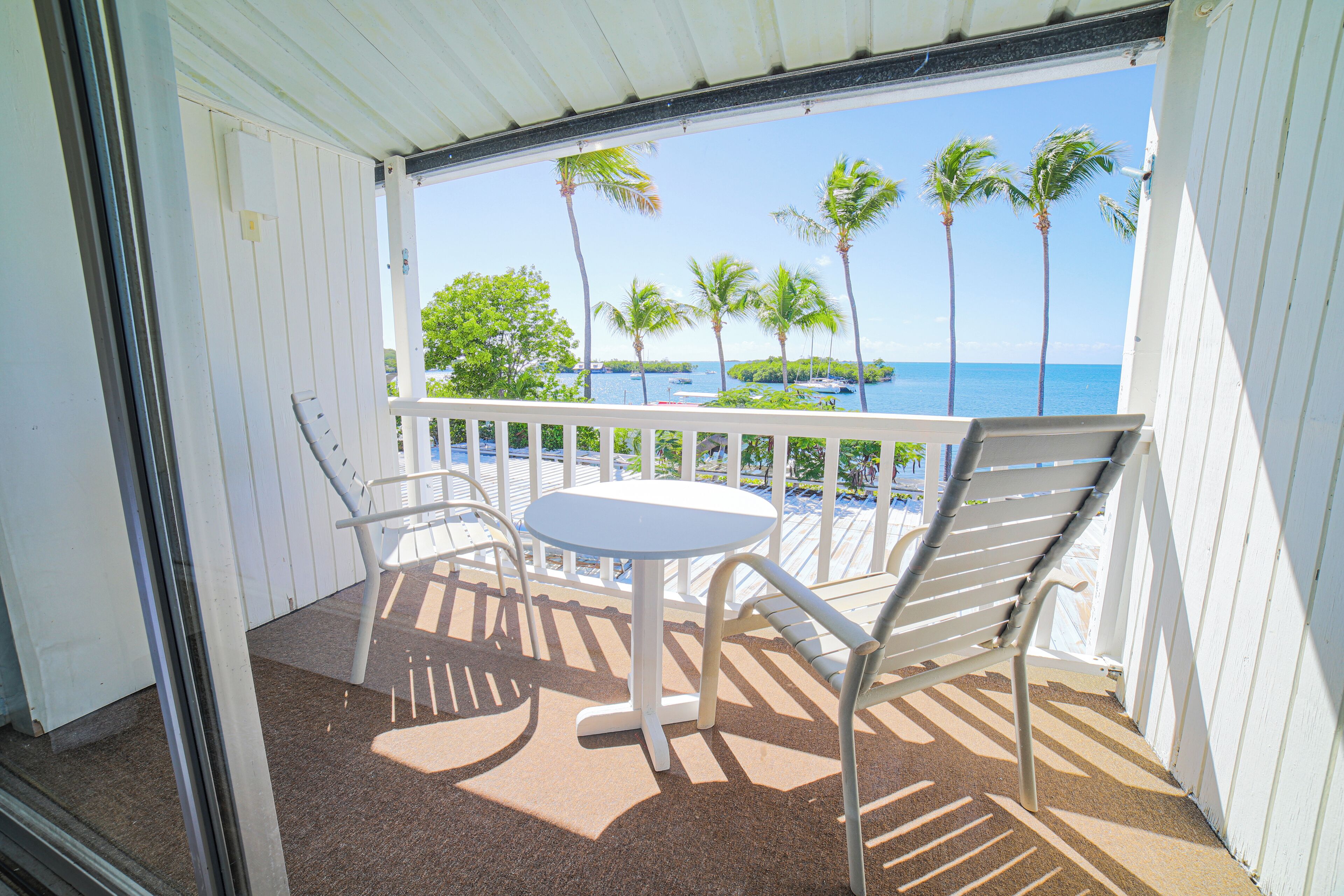 A balcony with a white table and two chairs, overlooking a beach with palm trees and boats.