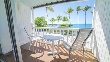 A balcony with a white table and two chairs, overlooking a beach with palm trees and boats.