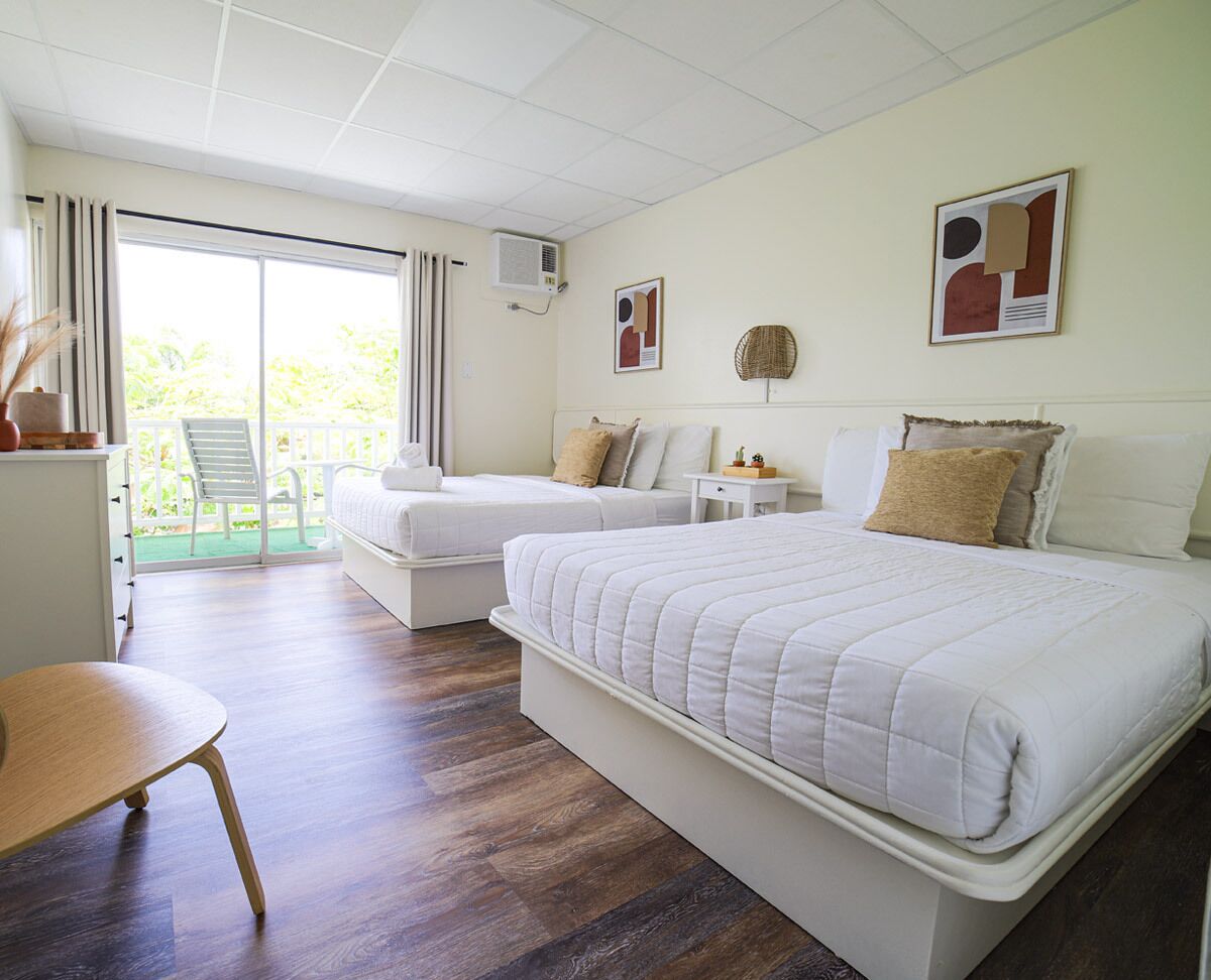 A hotel room with a large bed, two smaller beds, a desk, and a chair. There is a balcony with a view of greenery.