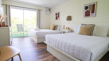 A hotel room with a large bed, two smaller beds, a desk, and a chair. There is a balcony with a view of greenery.