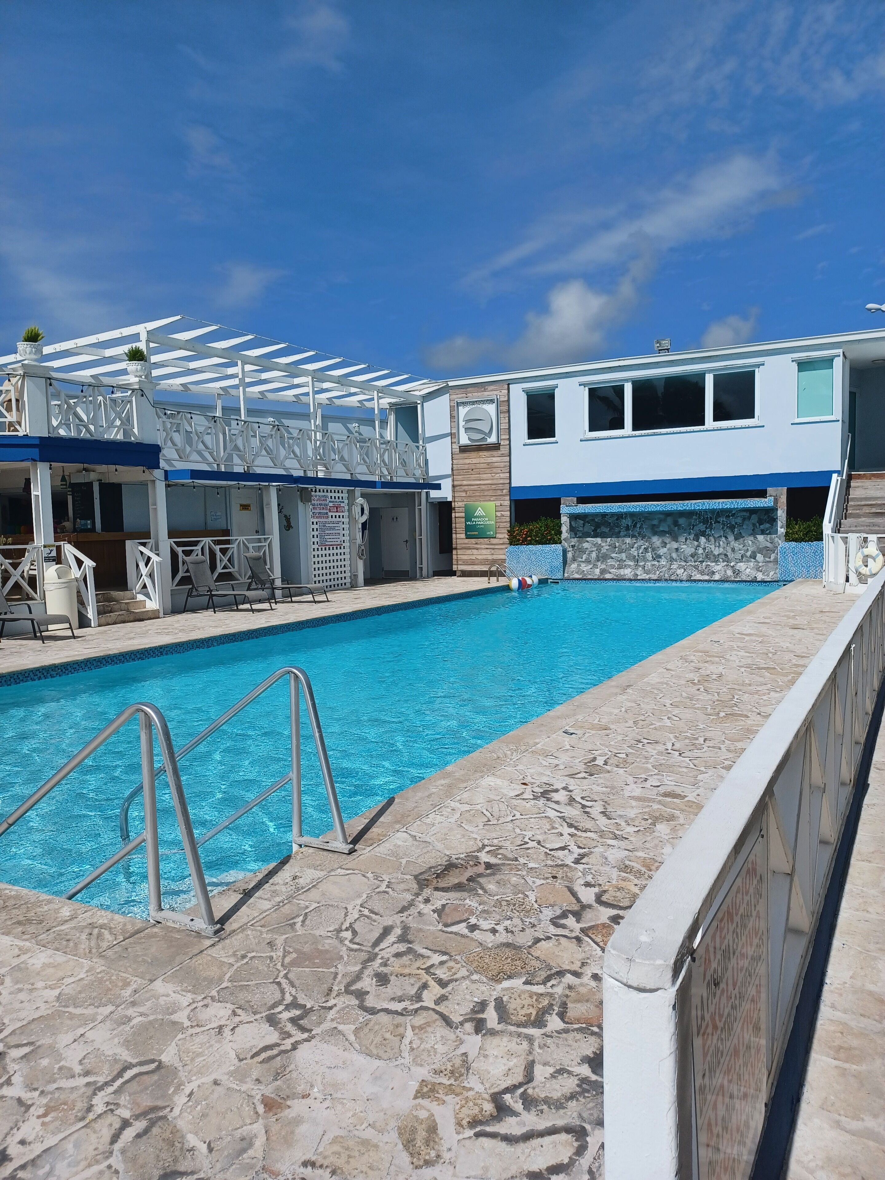 Una piscina con solárium de piedra, una zona de estar cubierta y un edificio de fachada azul y blanca.