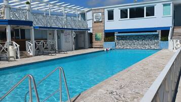 Una piscina con solárium de piedra, una zona de estar cubierta y un edificio de fachada azul y blanca.