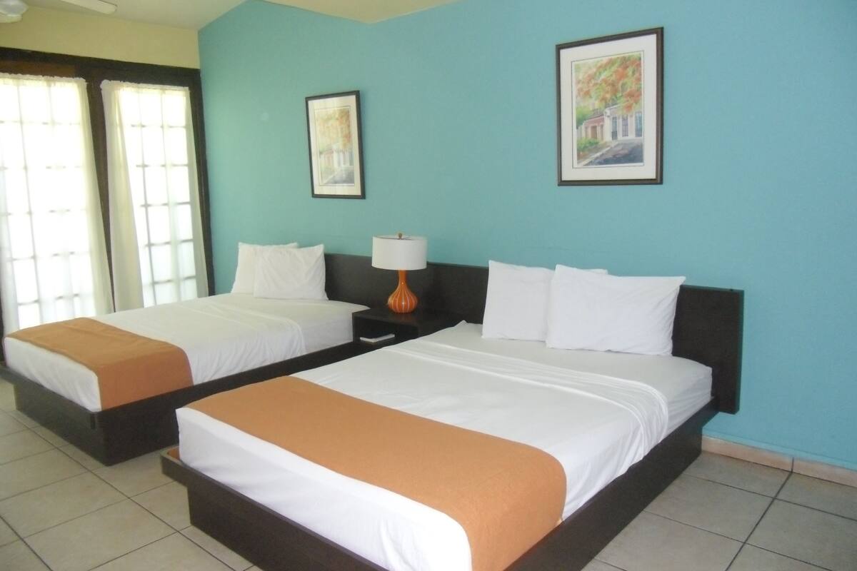 Quarto de hotel com duas camas, um ventilador de teto e duas quadros nas paredes.