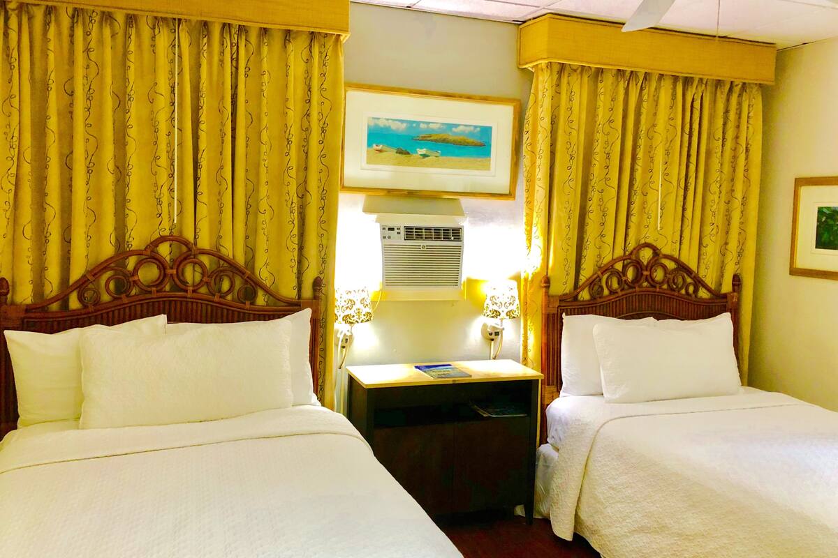 A hotel room with two beds, yellow curtains, a framed picture on the wall, and a bedside table with a lamp.