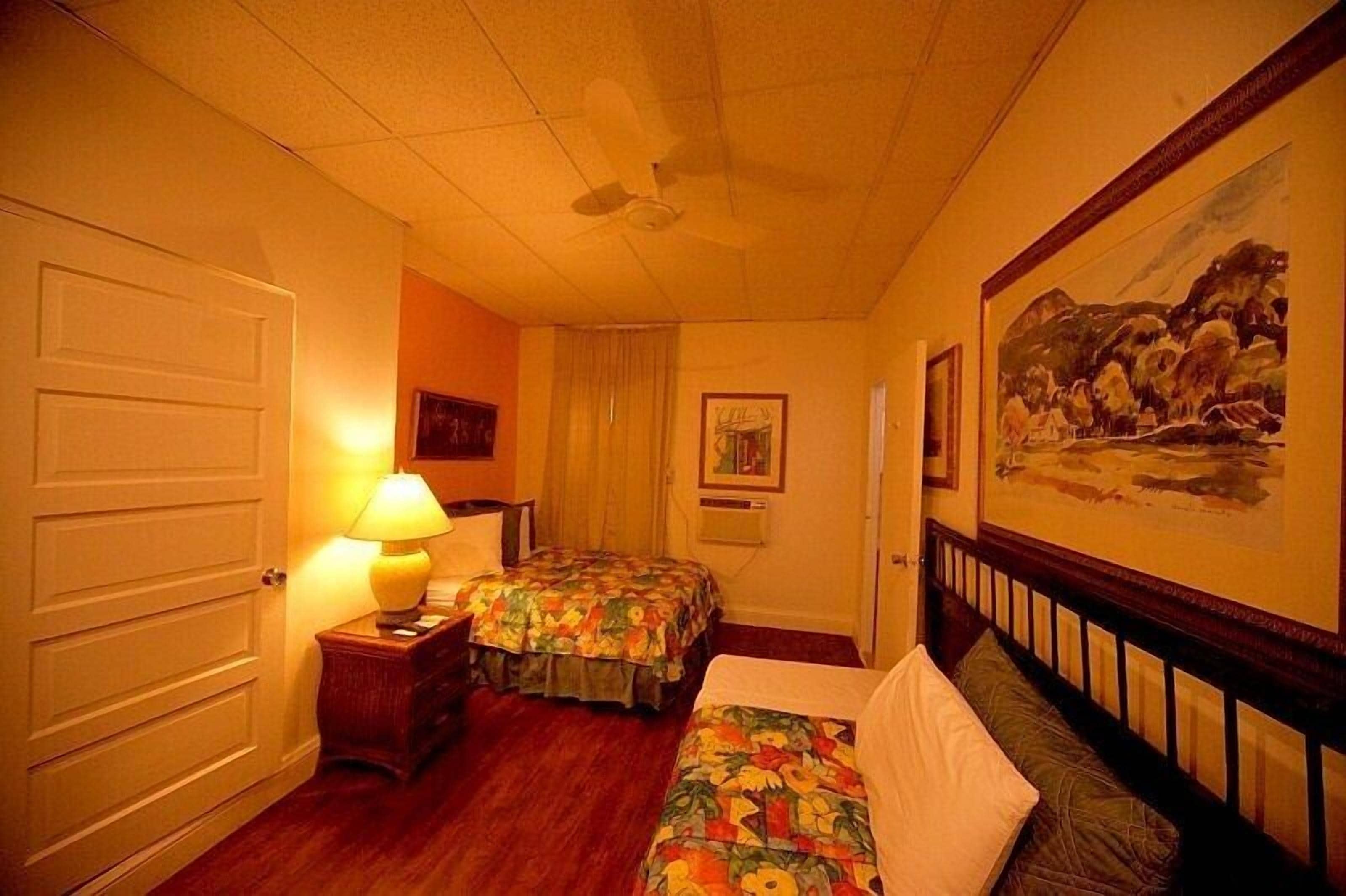 A hotel room with two beds, a ceiling fan, a painting on the wall, and a door.