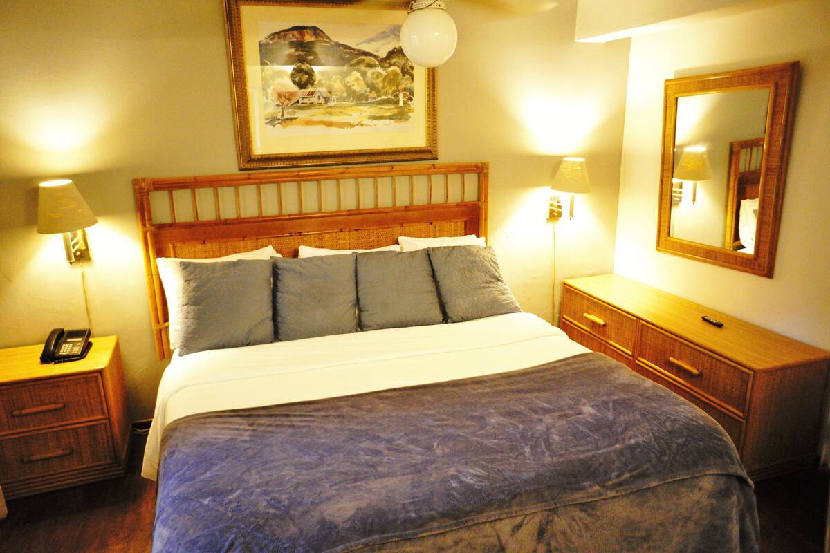 A hotel room with a wooden headboard, a large bed with multiple pillows, bedside lamps, a wooden nightstand with a phone, a dresser with drawers, and a framed picture on the wall.