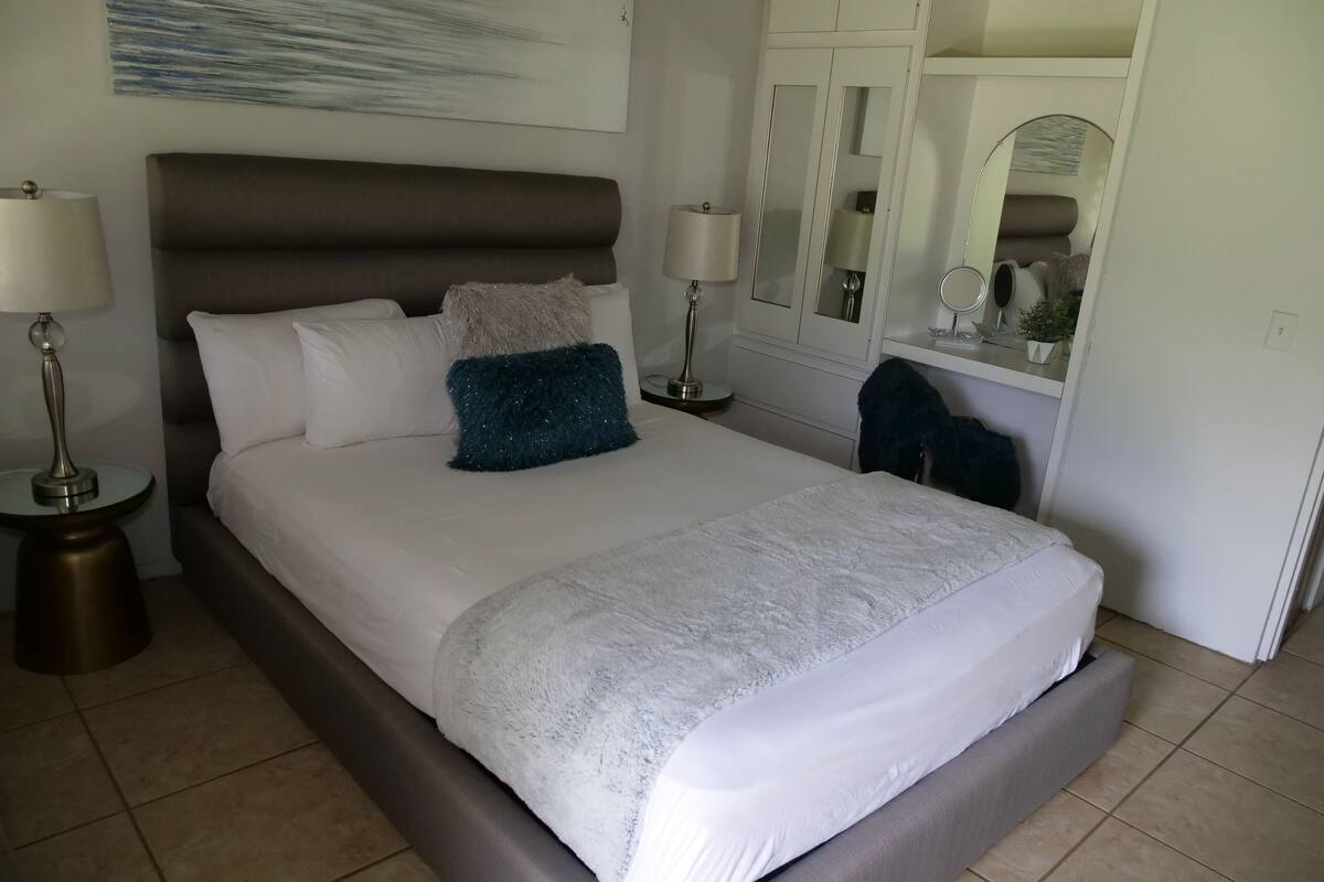Een keurig opgemaakt bed met witte lakens en een grijs hoofdbord, een bedlampje en een spiegel met een kleine plant op het nachtkastje.