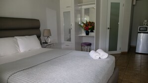 Una cama bien tendida con ropa de cama blanca, una lámpara de noche, un taburete con un jarrón de flores y un refrigerador en la esquina.