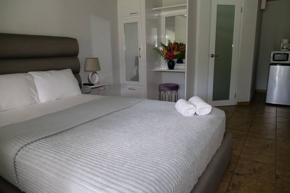 Een net opgemaakt bed met witte lakens, een bedlampje, een klein krukje met een bloemenvaas, en een koelkast in de hoek.