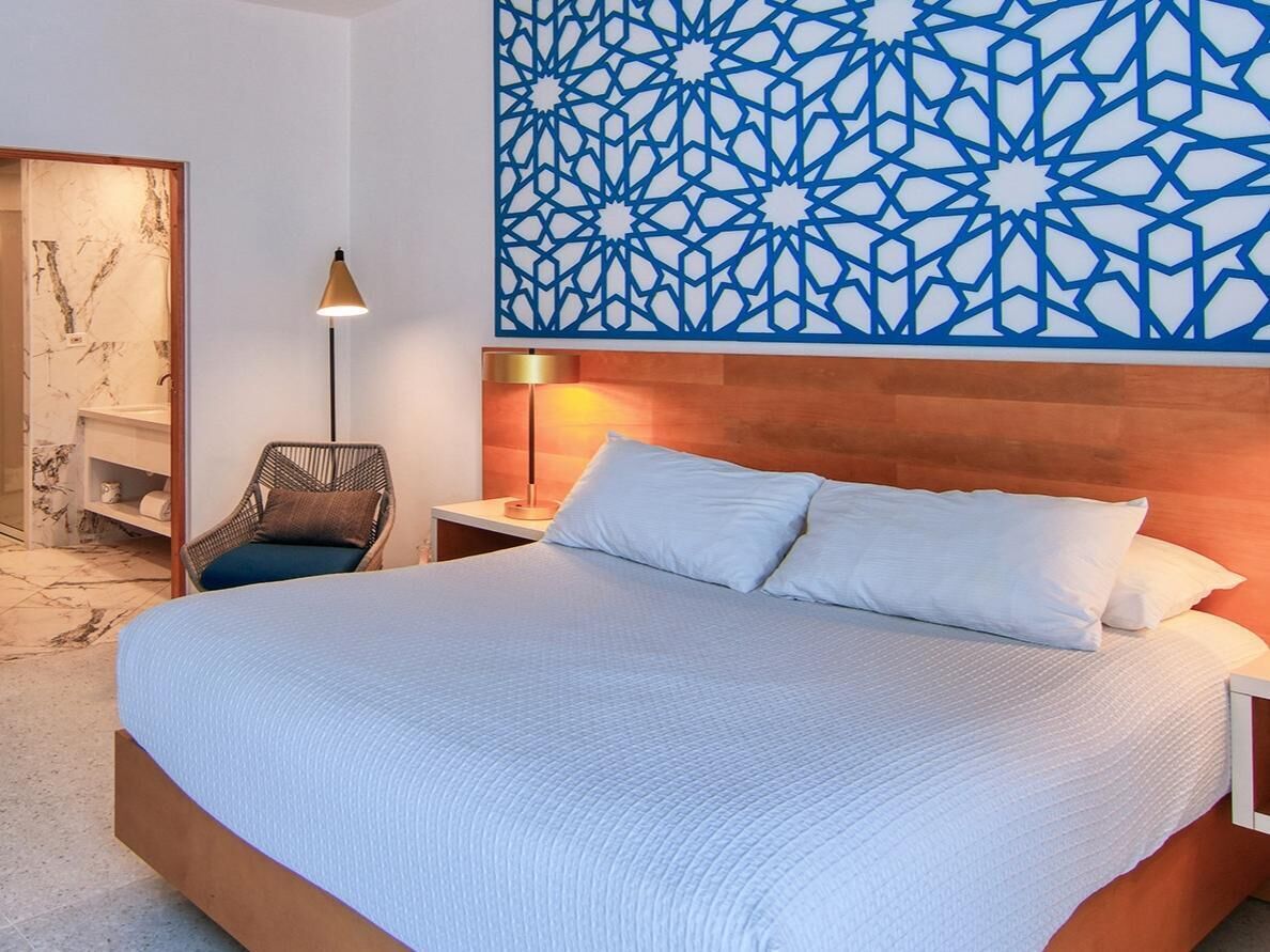 A hotel room with a large bed, a nightstand, a wall-mounted lamp, and a decorative geometric wall panel.