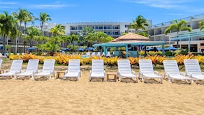 A beach with white lounge chairs and a pool area with umbrellas. - Rincon of the Seas - Grand Caribbean Hotel (Rincon)