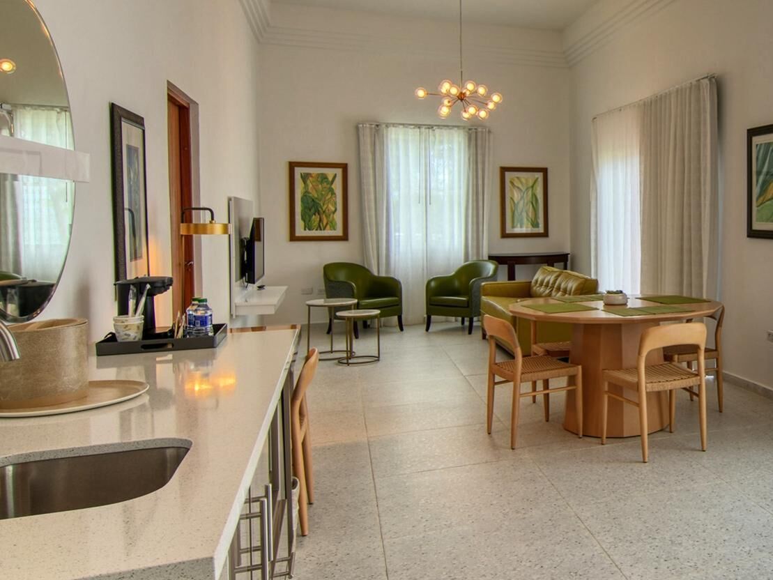A modern hotel room with a kitchen area, dining space, and a living area with green chairs and a round table.