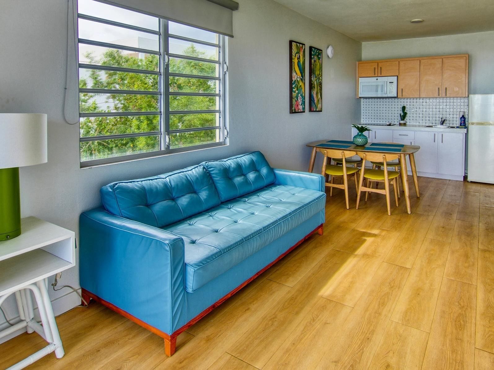 A modern living room with a blue sofa, wooden dining table, and a kitchen area with wooden cabinets and a white refrigerator.