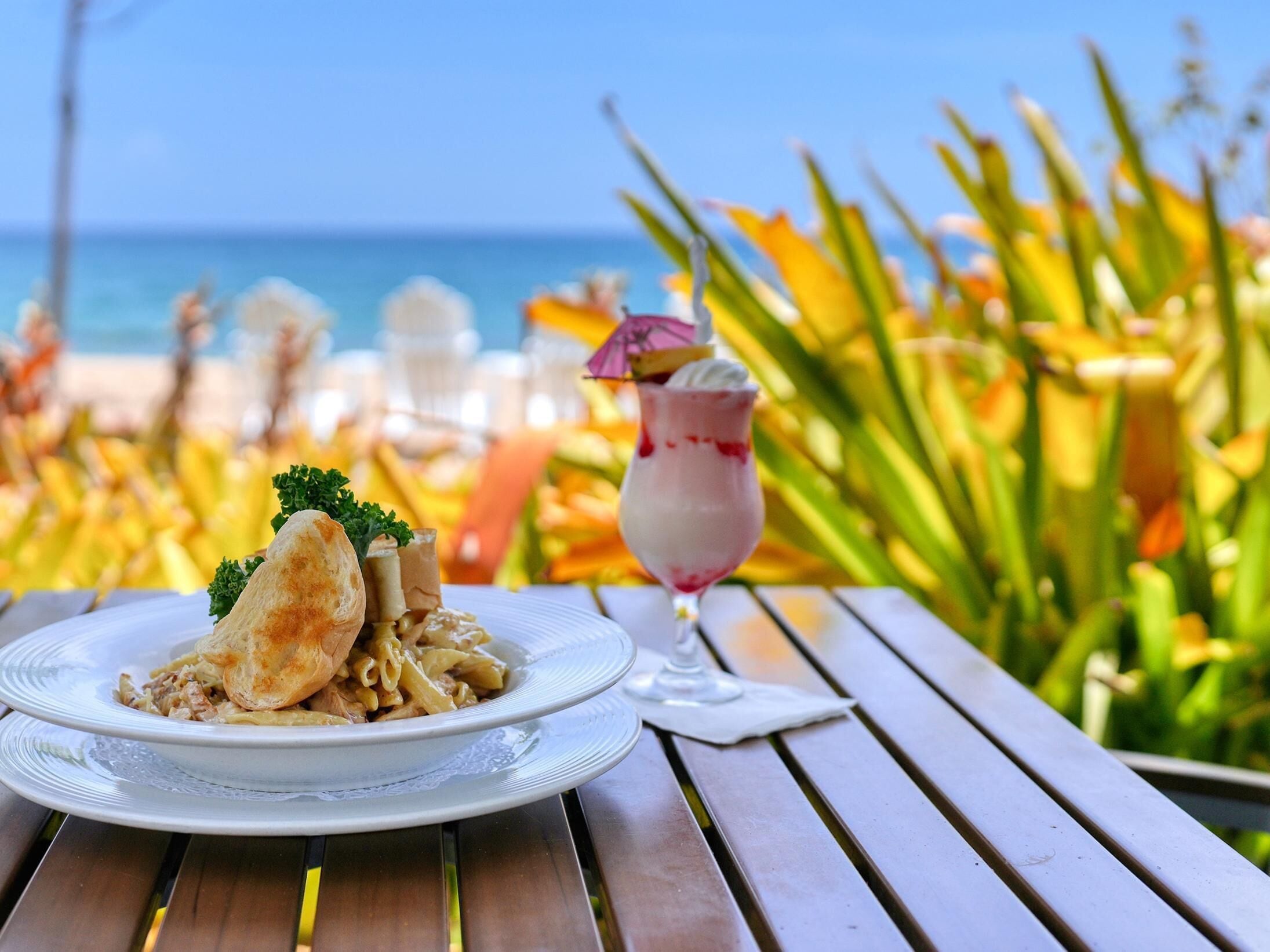 A plate of pasta with a bread roll and a glass of dessert on a wooden table with a view of the ocean and tropical plants.