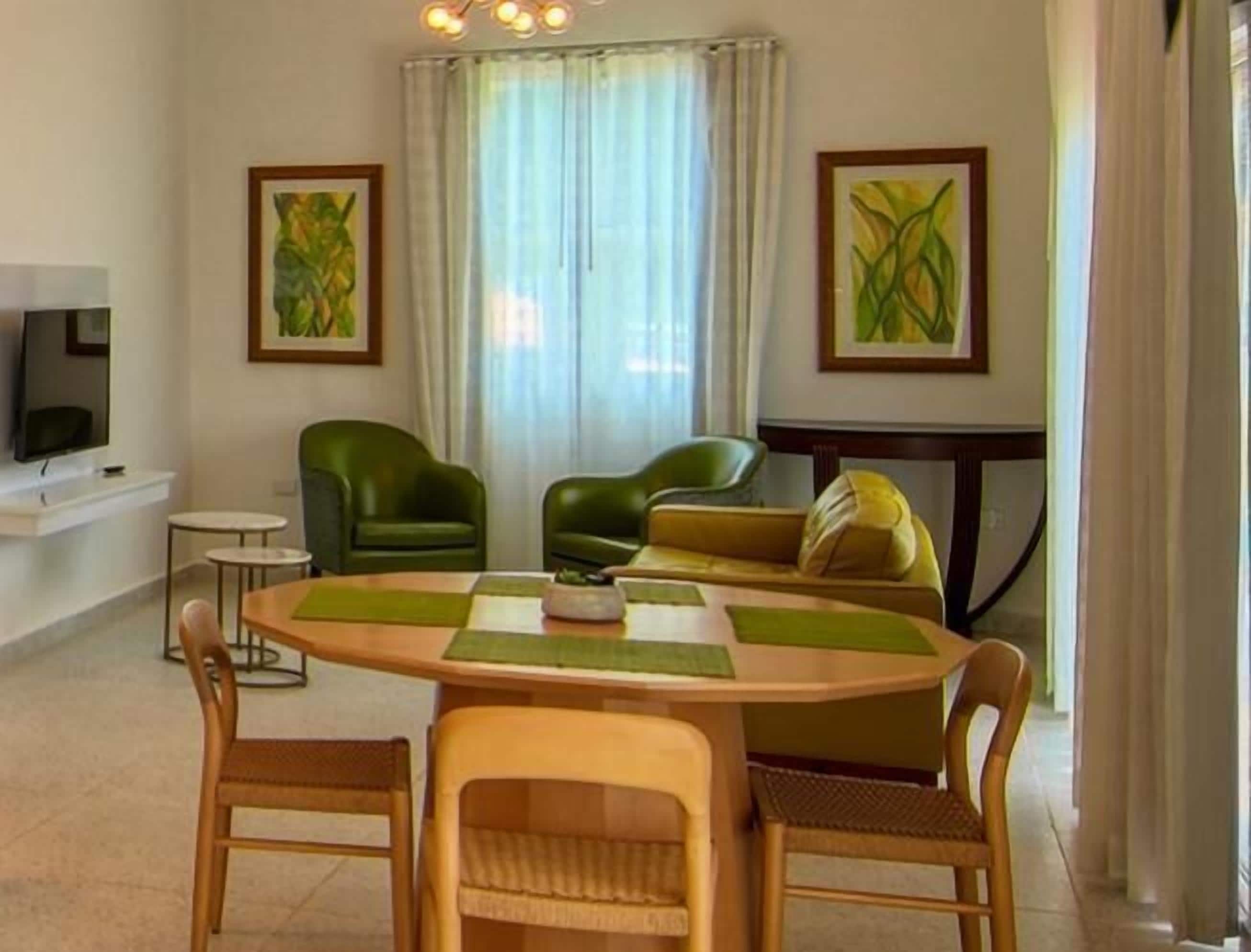 Ein Wohnzimmer mit Holztisch, grünen Stühlen, einer Couch und zwei Bildern an den Wänden.