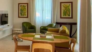 A living room with a wooden table, green chairs, a sofa, and two paintings on the walls.