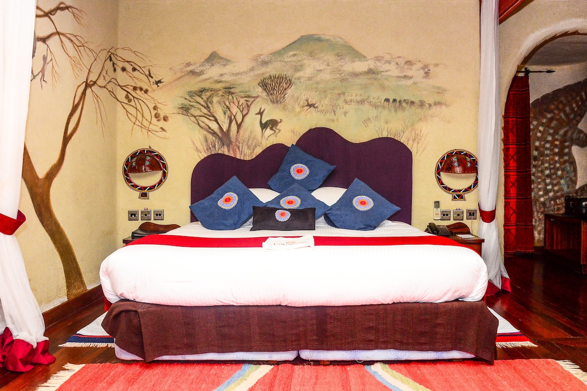 A bed with a white and brown bedspread, blue pillows with red and white patterns, and a mural of a landscape on the wall behind the bed.