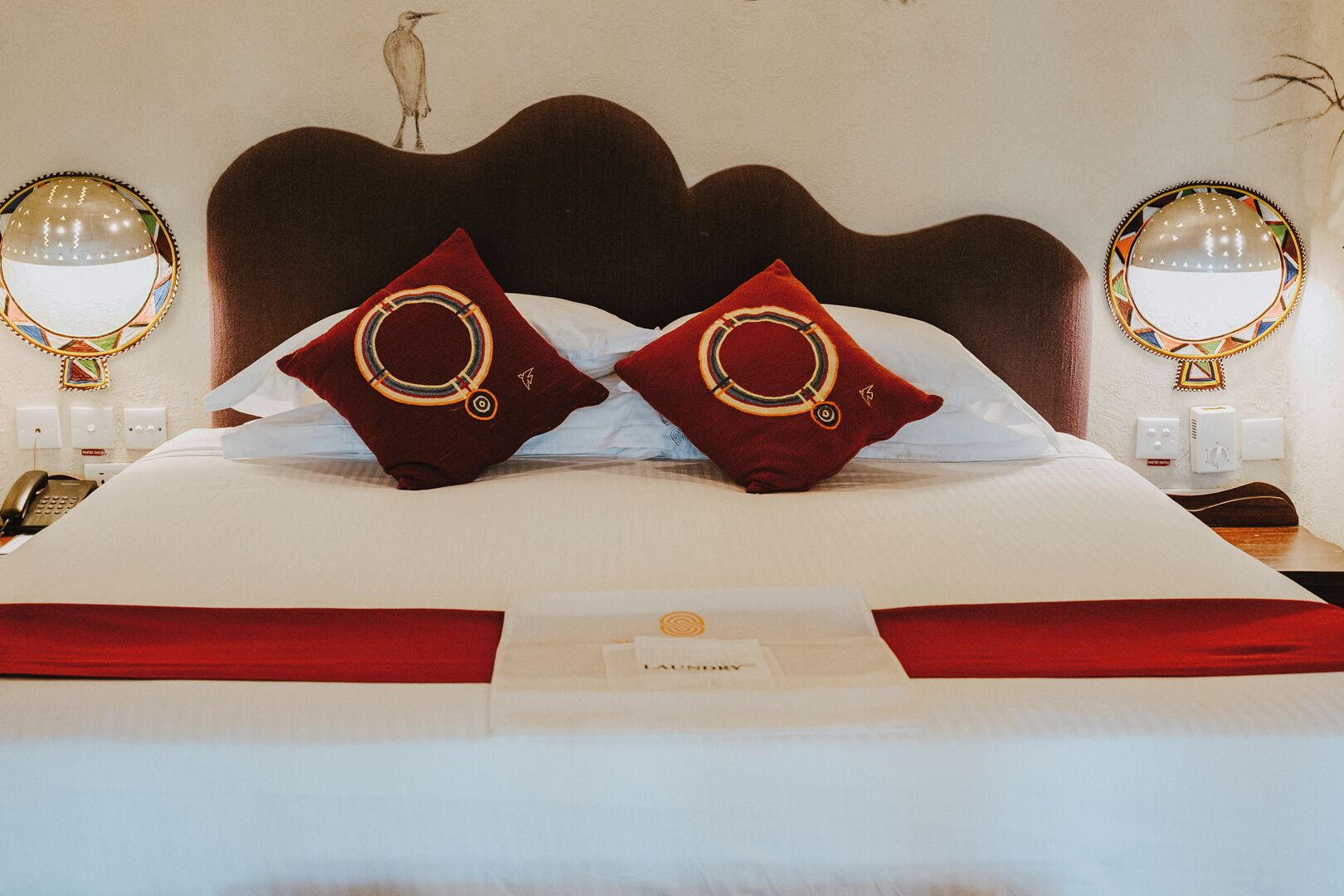 A hotel room with a large bed, two red pillows with a circular design, a wooden headboard, bedside lamps, a telephone, and a wall-mounted light fixture.