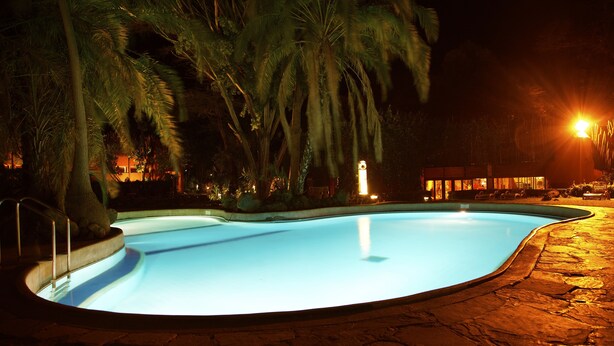 A nighttime swimming pool with a stone deck, surrounded by palm trees and a building with illuminated windows in the background. - Amboseli Serena Safari Lodge (Amboseli)