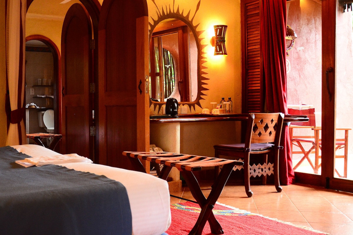 A hotel room with a bed, a desk with a chair, a red rug, and a view of the outside.