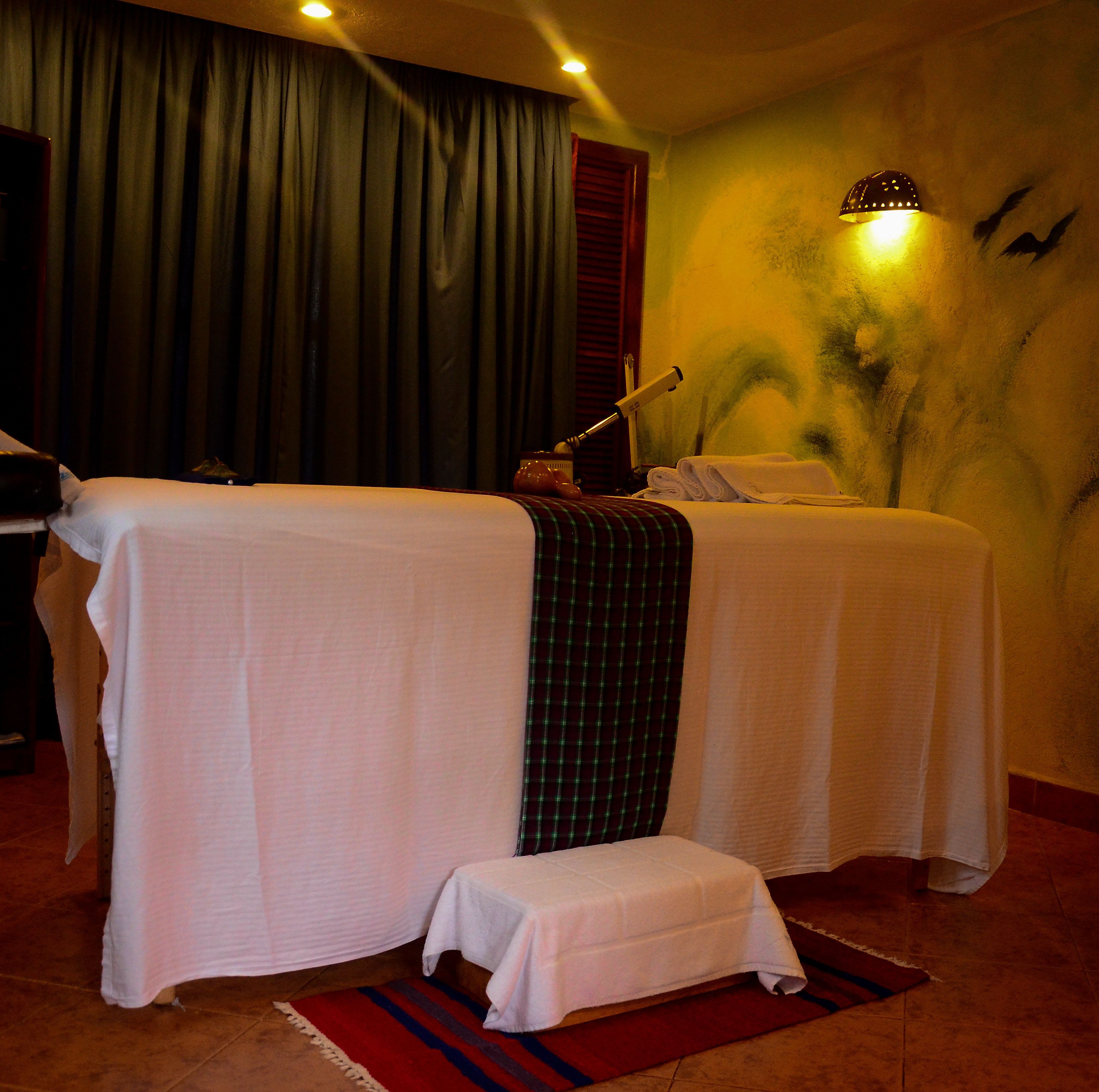 A room with two massage tables covered in white sheets, a small bench, a wall-mounted lamp, and a curtain.