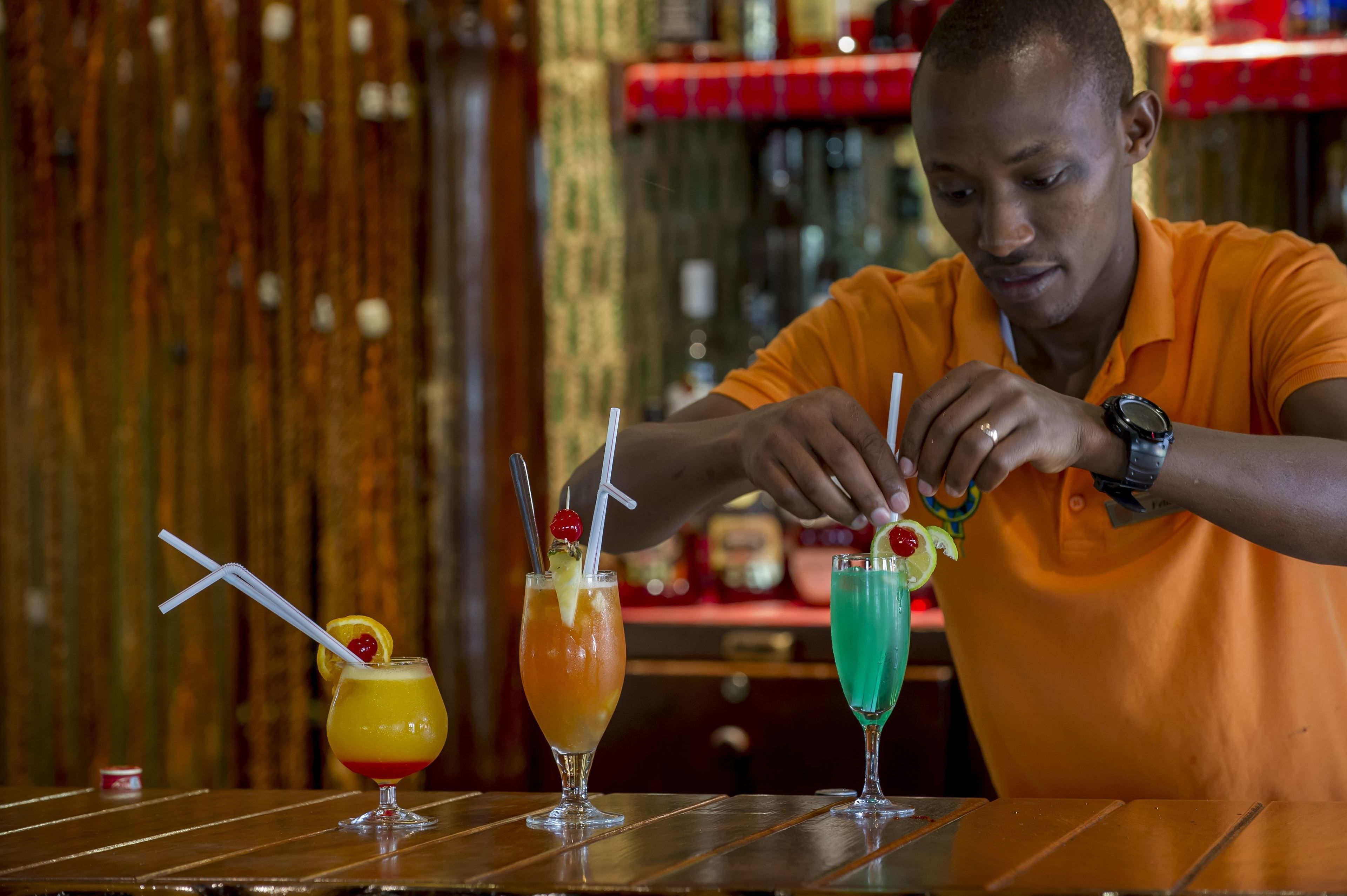 Une personne portant un t-shirt orange prépare des boissons dans un bar dont la décoration est en bois et où la table est recouverte d’une nappe à carreaux rouges.