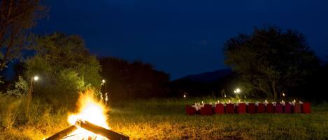 Un feu de camp dans un champ herbeux, avec des tables et des chaises disposées pour les repas, entouré d’arbres et sous un ciel sombre.