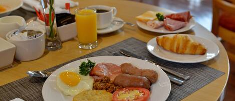 Una mesa de desayuno con platos de huevos, salchichas y tomates, un vaso de zumo de naranja y un croissant.