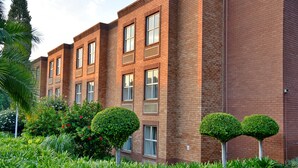 Un edificio de varios pisos de ladrillo con fachada simétrica, ventanas amplias y un jardín bien cuidado con arbustos podados y una palmera.