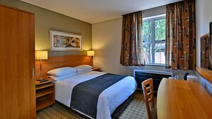 Una habitación de hotel con cama, mesita de noche, lámpara y ventana con cortinas.