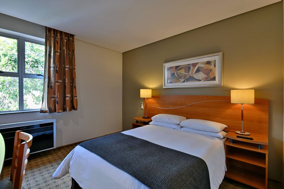 Habitación de hotel con cama, dos lámparas de noche, cabecero, cuadro abstracto enmarco, ventana con cortinas, radiador y una silla.