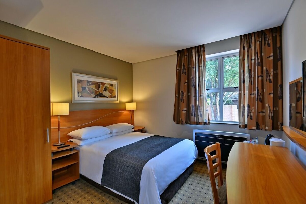 Een hotelkamer met een bed, nachtkastje, lamp, stoel en een raam met gordijnen.